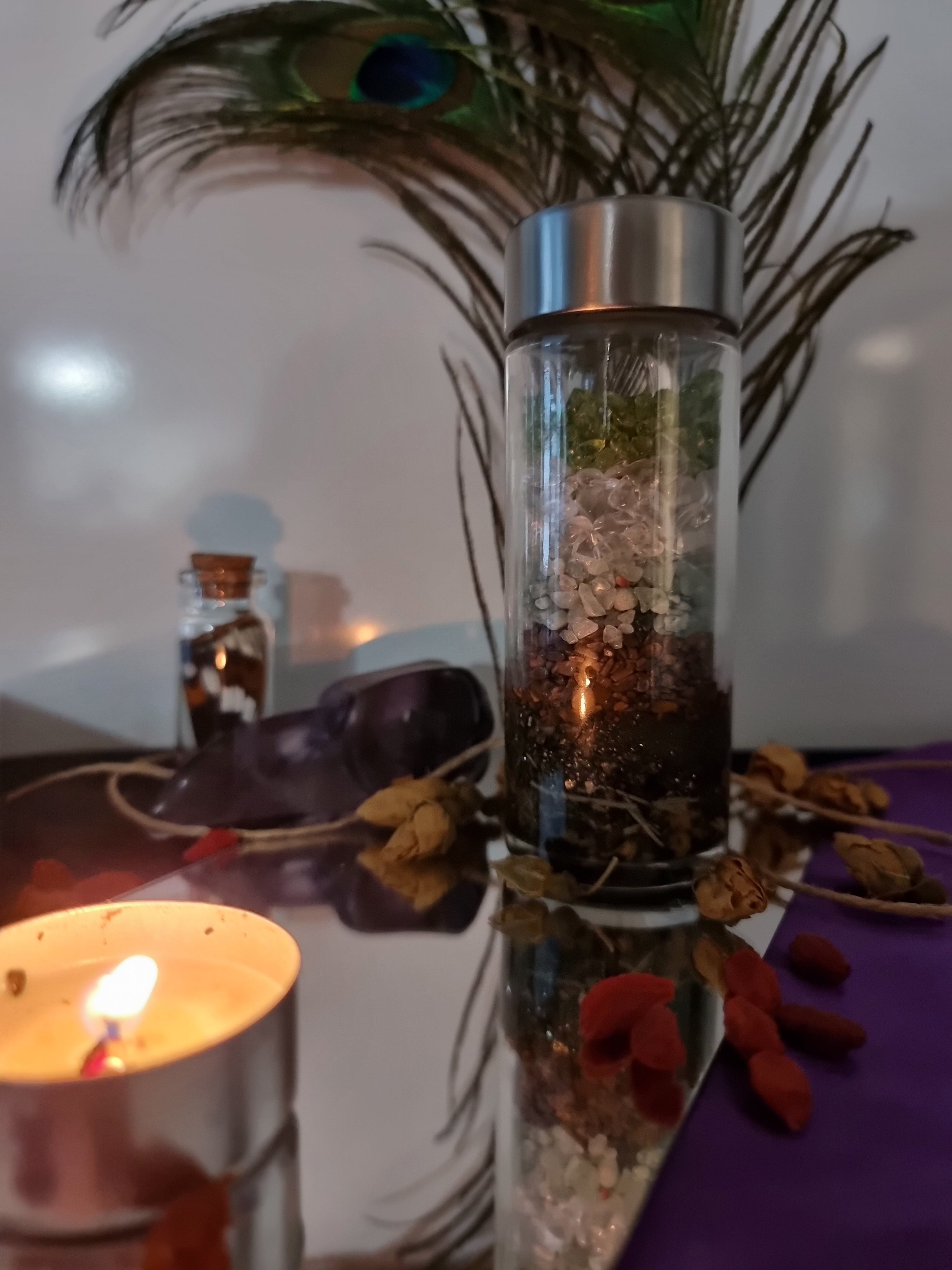 Decorative glass bottle with peacock feather, flowers, and a lit candle on a reflective surface.